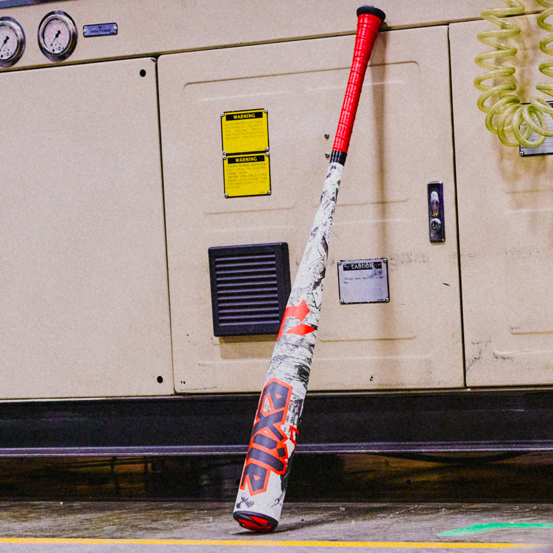 A white DeMarini 2026 Exile (-3) BBCOR Baseball Bat (WBD2589010) with red and black accents leans against an industrial machine with gauges, vents, yellow warning labels, and a coiled air hose hanging to the right.