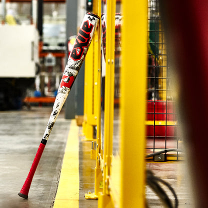 A 2026 DeMarini Exile (-3) BBCOR Baseball Bat (WBD2589010) by DeMarini, featuring a red grip and graphic design, leans against a yellow metal fence in an industrial warehouse setting.
