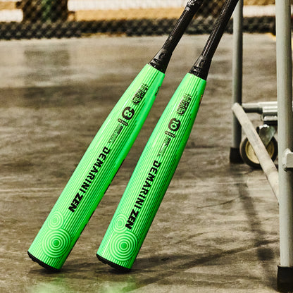 Two bright green 2026 DeMarini ZEN (-8) 2 3/4" USSSA Baseball Bats (WBD2592010), each with two-piece construction and black handles, stand upright against a metal fence on a concrete floor, showing brand and model details in black text.