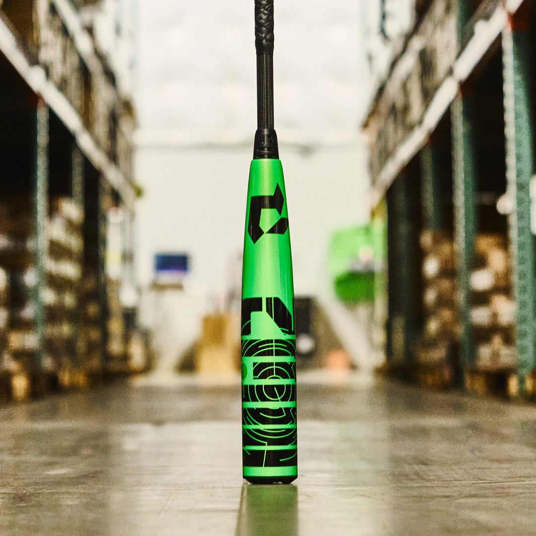 A green and black DeMarini 2026 ZEN (-8) 2 3/4" USSSA Baseball Bat (WBD2592010), featuring two-piece construction, stands upright on the warehouse floor with shelves of boxes blurred in the background.