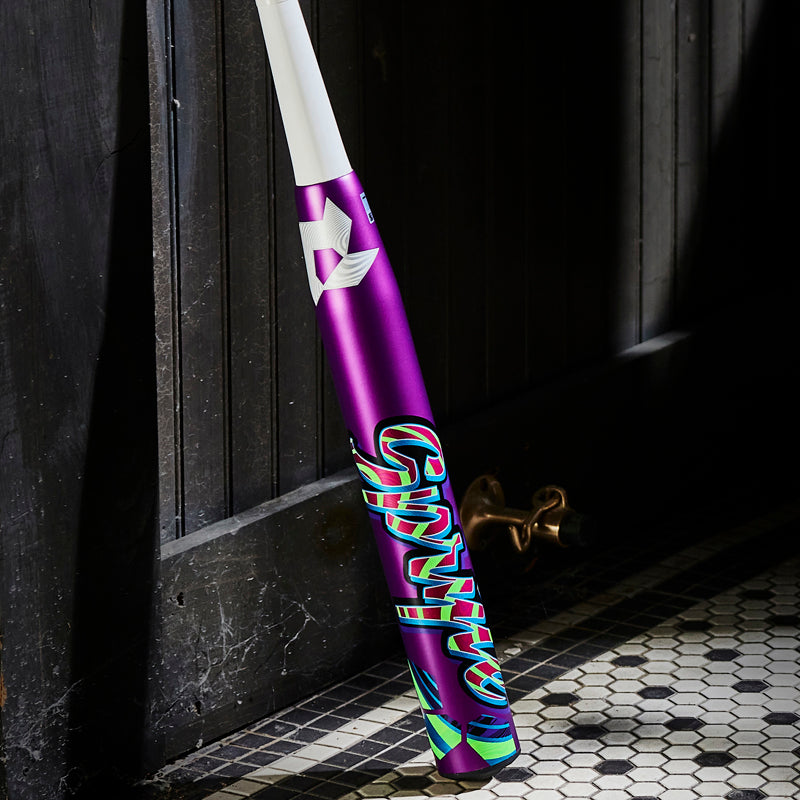 A 2025 DeMarini Spryte (-12) Fastpitch Softball Bat (WBD2553010) with purple and white graphics leans against a dark wooden door on a patterned tile floor.