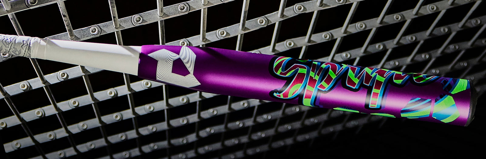 A colorful purple baseball bat with white grip tape rests against a metallic chain-link fence under bright lighting.