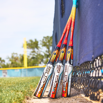 Three colorful baseball bats, including a 2026 Louisville Slugger Supra Starship (-10) USSSA Baseball Bat, lean against a blue fence on a field with green grass, a dirt path, and a yellow outfield fence in the background.