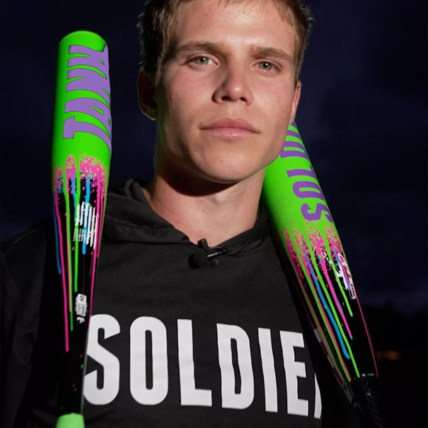 A young man in a black hoodie with SOLDIER in white holds two neon green Soldier Sports 2026 Soldier Tank2 (-3) BBCOR Baseball Bats (BB-1-26) over his shoulders against a dark, cloudy evening sky.