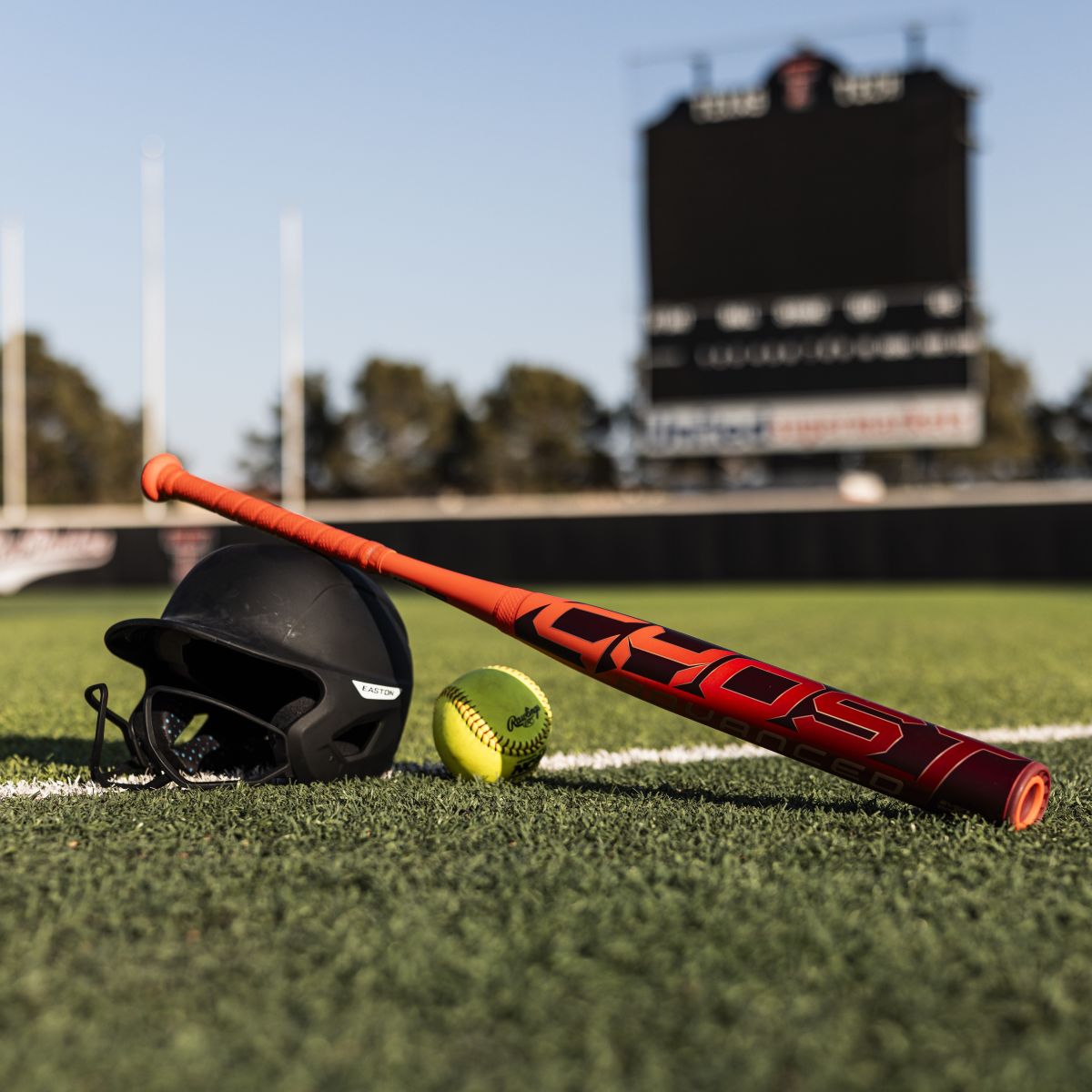 A black helmet, yellow softball, and 2026 Easton Ghost Advanced (-8) Fastpitch Softball Bat by Easton rest on artificial turf in front of a blurred stadium scoreboard.