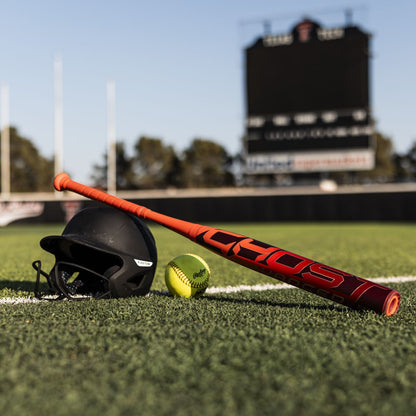 A black helmet, yellow softball, and 2026 Easton Ghost Advanced (-8) Fastpitch Softball Bat by Easton rest on artificial turf in front of a blurred stadium scoreboard.