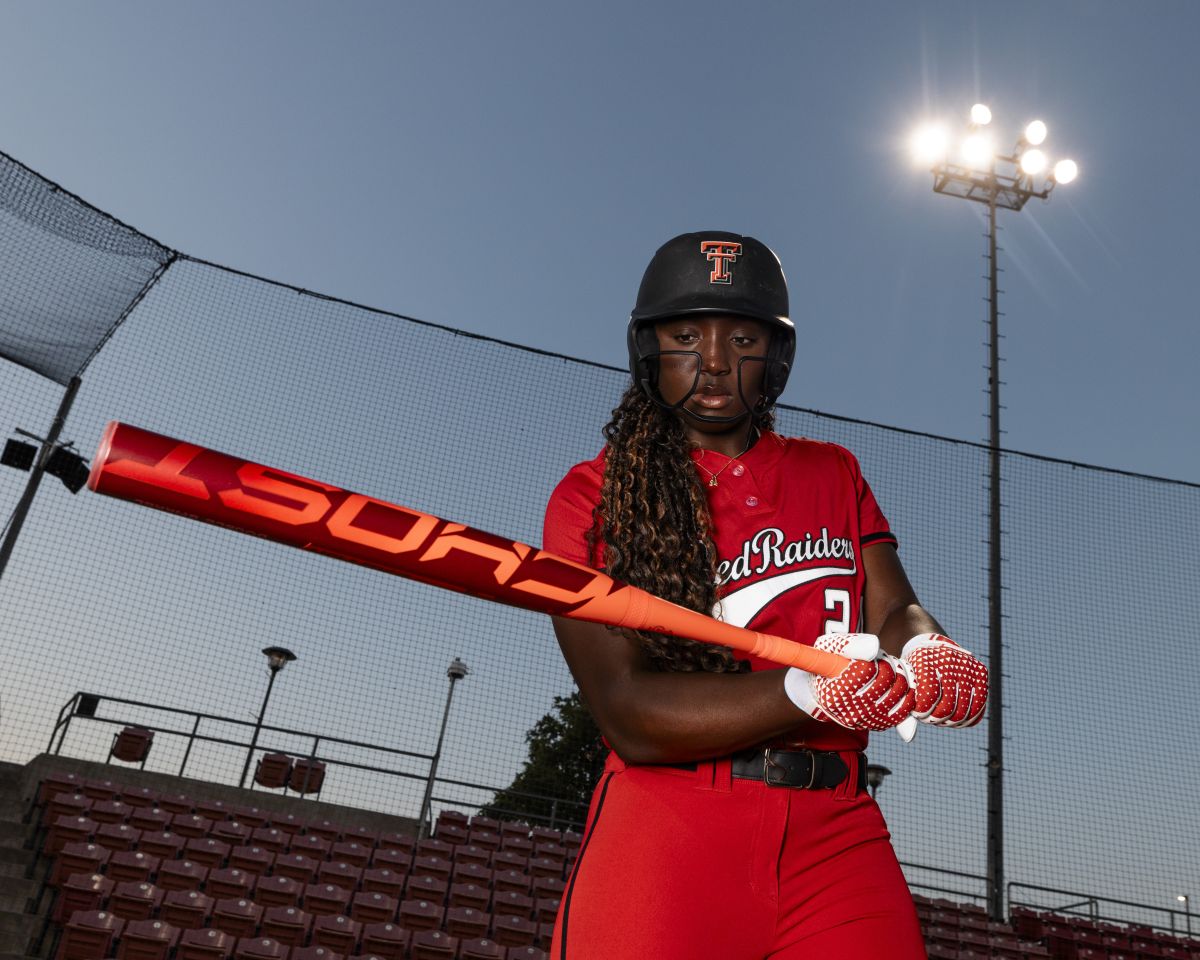 A softball player in a red uniform and helmet stands ready to swing the 2026 Easton Ghost Advanced (-8) Fastpitch Softball Bat (EFP6GHAD8) by Easton under bright stadium lights, with empty bleachers in the background at dusk.