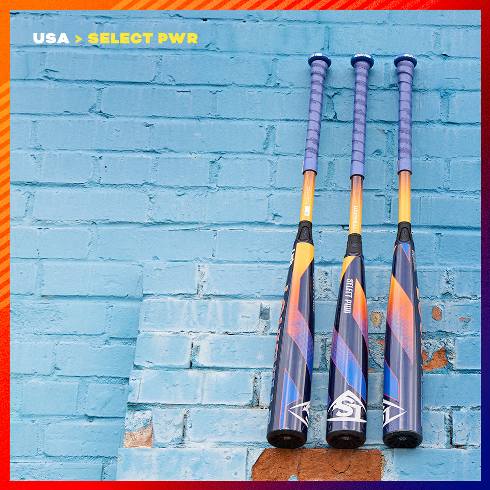 Three baseball bats with blue and orange designs and purple grips are leaning upright against a light blue brick wall. The text USA > SELECT PWR appears in the top left corner.