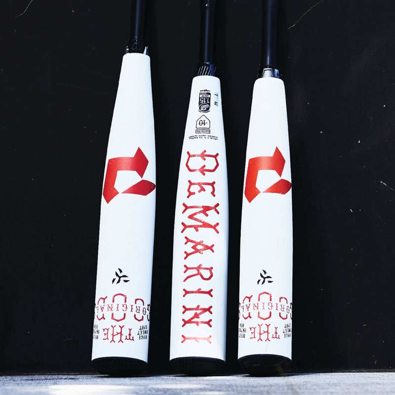 Three white baseball bats with black handles are standing upright, featuring red and black text and graphics, including the word DEMARINI printed vertically in bold red letters on the center bat.