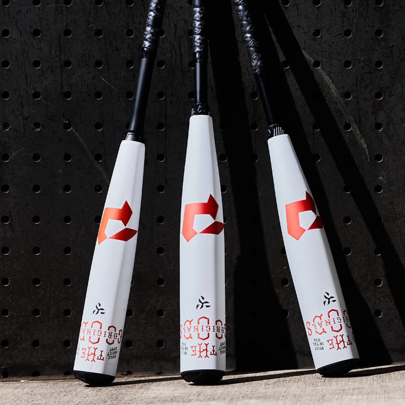 Three white baseball bats with red logos and black handles are standing upright against a dark perforated background, with sunlight casting shadows on the ground.