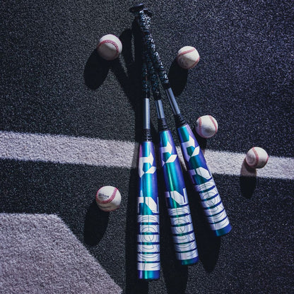 Three DeMarini 2026 ZEN Oil Slick (-10) USSSA Baseball Bats (WBD2655010) with geometric patterns and a metallic blue finish are lined up on asphalt beside five baseballs, dramatic shadows and light enhancing their vibrant look.