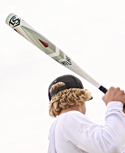 A person with blonde hair in a black mesh cap and white shirt holds the 2025 Louisville Slugger Atlas (-5) 2 5/8" USSSA Baseball Bat (WBL2981010) over their shoulder, ready to swing, photographed from behind against a bright, cloudy sky.