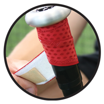 Close-up of a hand wrapping a red grip tape around the handle of a black tennis racket, with a blurred green background.