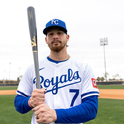A baseball player in a white Royals jersey and blue cap stands on the field, holding a Victus Bobby Witt Jr BWITT7 Pro Reserve Maple Wood Bat (VRWMBWITT7) vertically. Behind him is an empty diamond with light poles under a cloudy sky.