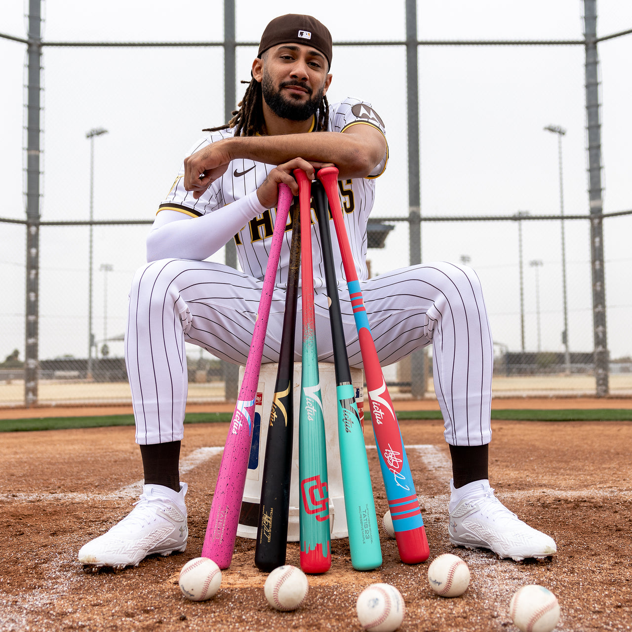 Wearing a striped uniform and white sneakers, a baseball player sits on home plate with colorful bats—including the Victus TATIS JR Youth Pro Reserve Birch Wood Bat—and enjoys its balanced feel, several baseballs at his feet and a chain-link fence in the background.