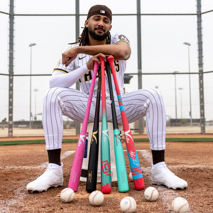 Wearing a striped uniform and white sneakers, a baseball player sits on home plate with colorful bats—including the Victus TATIS JR Youth Pro Reserve Birch Wood Bat—and enjoys its balanced feel, several baseballs at his feet and a chain-link fence in the background.