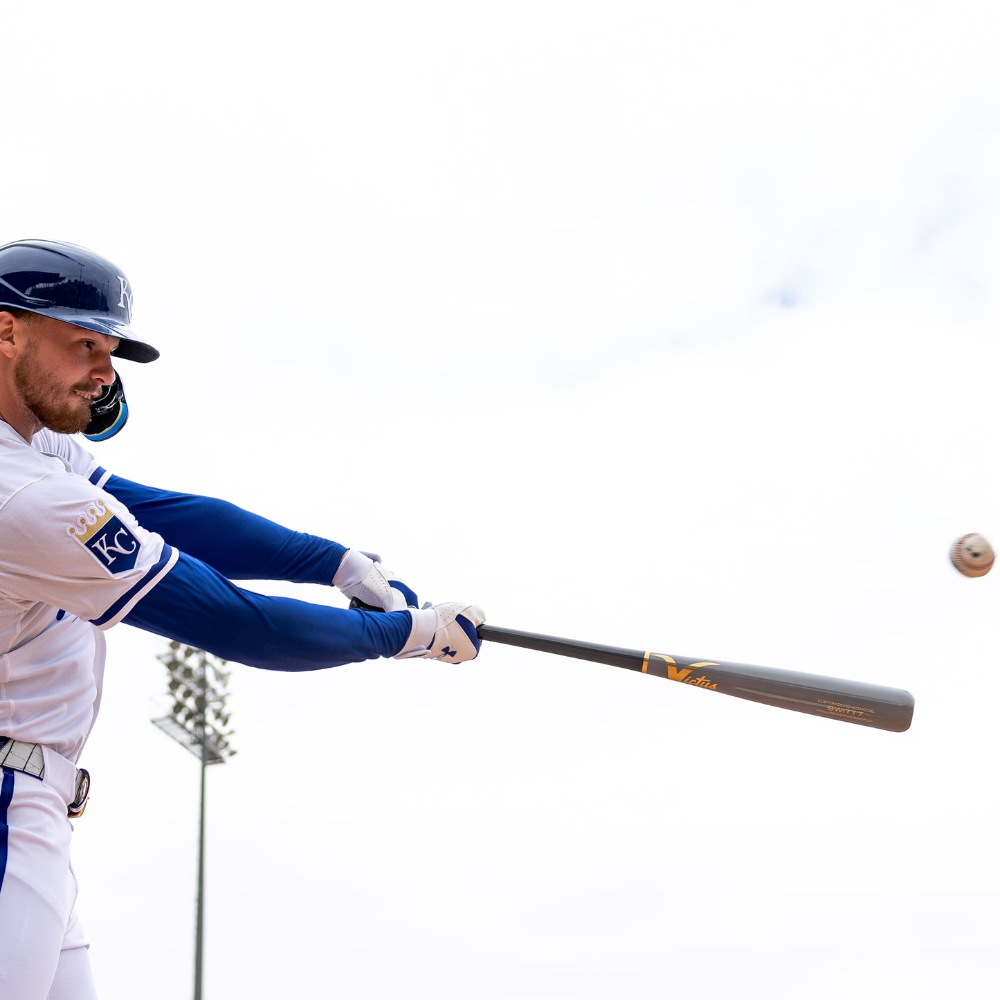 A baseball player in a white and blue uniform swings a Victus Bobby Witt Jr BWITT7 Pro Reserve Maple Wood Bat (VRWMBWITT7) by Victus, making contact with the ball during a game on a bright, cloudy day with a stadium light pole in the background.