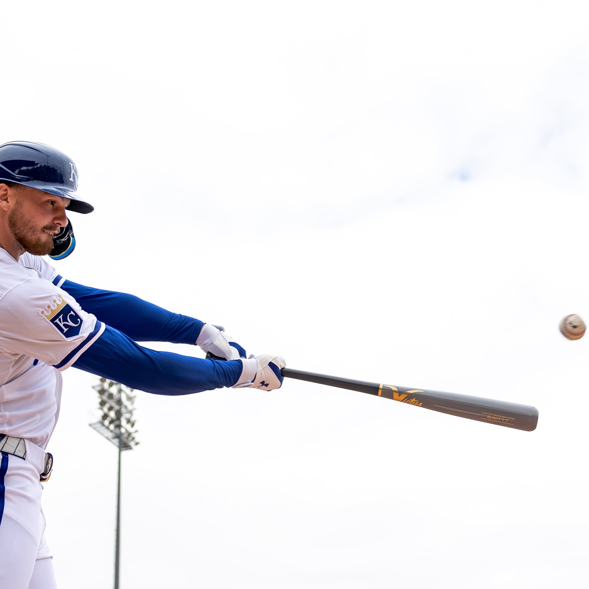 A baseball player in a white and blue uniform swings a Victus Bobby Witt Jr BWITT7 Pro Reserve Maple Wood Bat (VRWMBWITT7) by Victus, making contact with the ball during a game on a bright, cloudy day with a stadium light pole in the background.