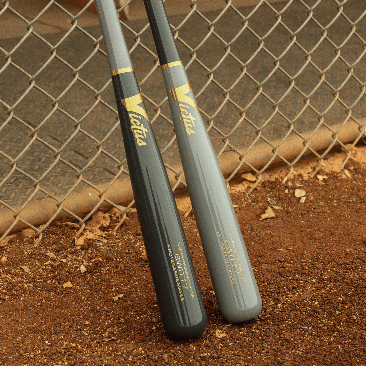 Two Victus Bobby Witt Jr BWITT7 Pro Reserve maple wood bats by Victus, one dark gray and one light gray, stand upright on a baseball field. Both VRWMBWITT7 bats feature gold branding against a chain-link fence.
