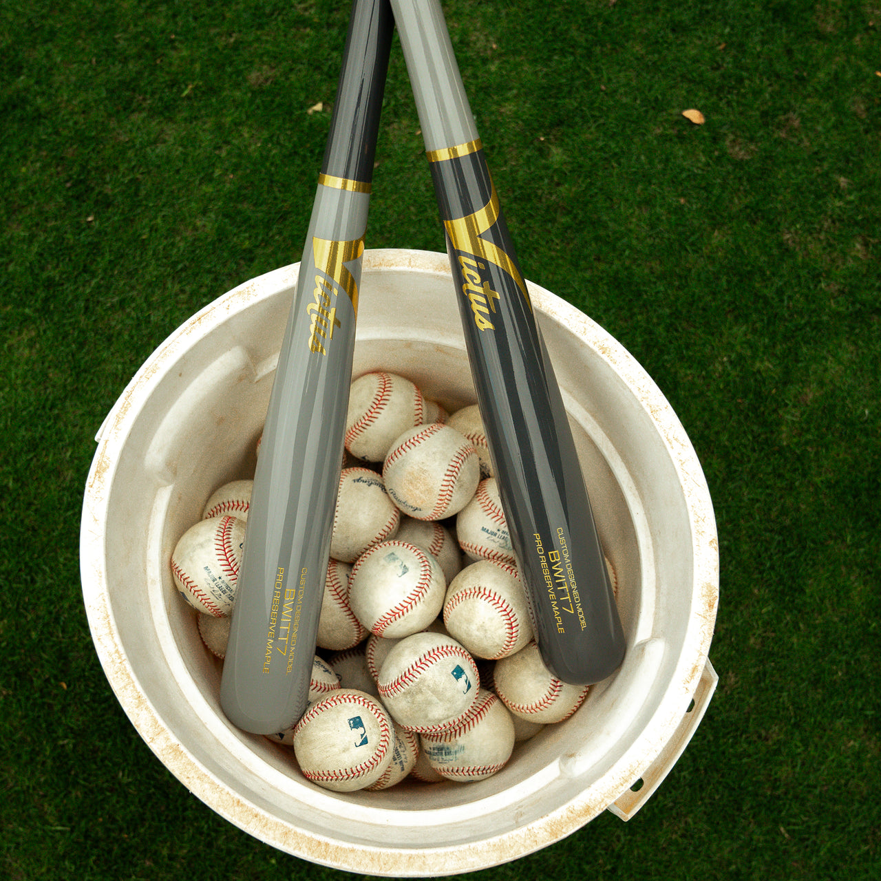 A white bucket filled with baseballs and two Victus Bobby Witt Jr BWITT7 Pro Reserve Maple Wood Bats (brand: Victus) resting across the top, placed on a grassy field.