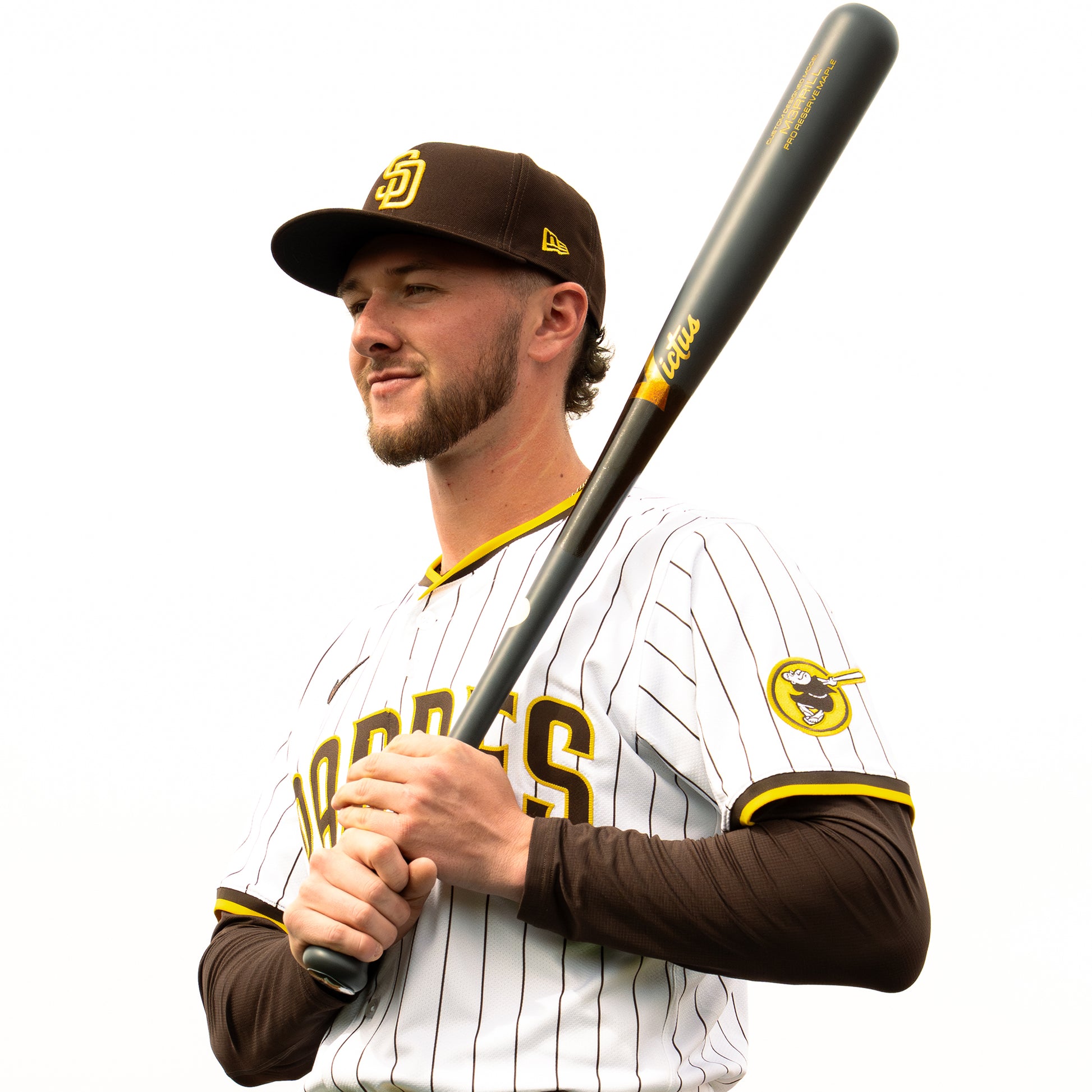A baseball player in a white pinstripe Padres uniform and brown cap stands holding a black Victus Jackson Merrill M3RRILL Pro Reserve Maple Wood Bat (VRWMM3RRILL), smiling and looking to the side against a white background.