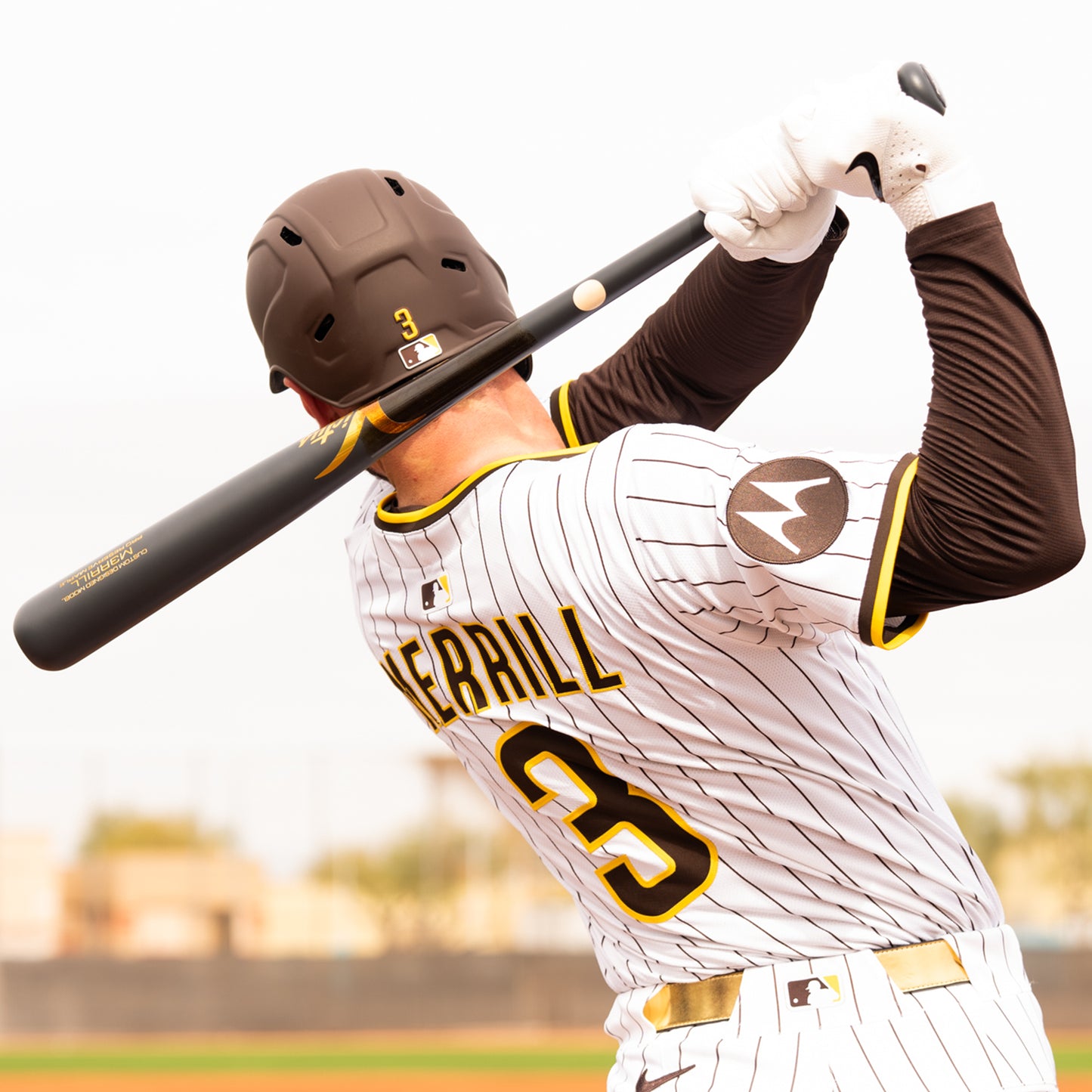 A baseball player in a white pinstripe uniform with "MERRILL" and number 3 uses a Victus Jackson Merrill M3RRILL Pro Reserve Maple Wood Bat (VRWMM3RRILL) during play, with a blurry field and trees visible behind him.