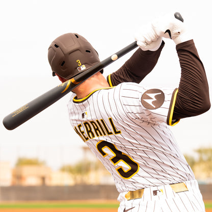 A baseball player in a white pinstripe uniform with "MERRILL" and number 3 uses a Victus Jackson Merrill M3RRILL Pro Reserve Maple Wood Bat (VRWMM3RRILL) during play, with a blurry field and trees visible behind him.