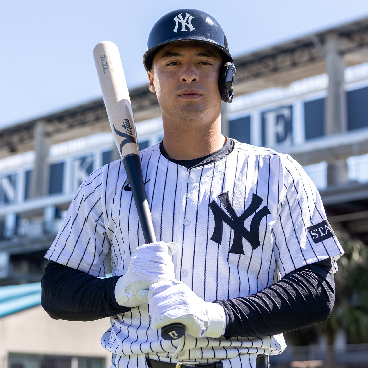 A baseball player in a New York Yankees uniform stands holding a Victus Anthony Volpe VOLPE1 Pro Reserve Maple Wood Bat (VRWMVOLP1), wearing a helmet and gloves, with a stadium structure in the background.