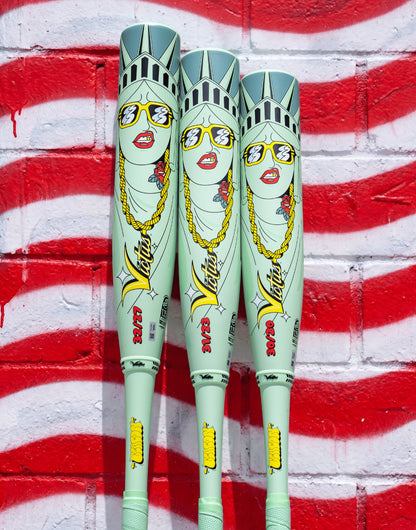 Three 2027 Victus Nova Lit Beacon (-10) USSSA Baseball Bats (VSBNL10B), featuring cartoon-style Statue of Liberty graphics with sunglasses, gold chains, and red lipstick, stand upright against a red and white brick wall.