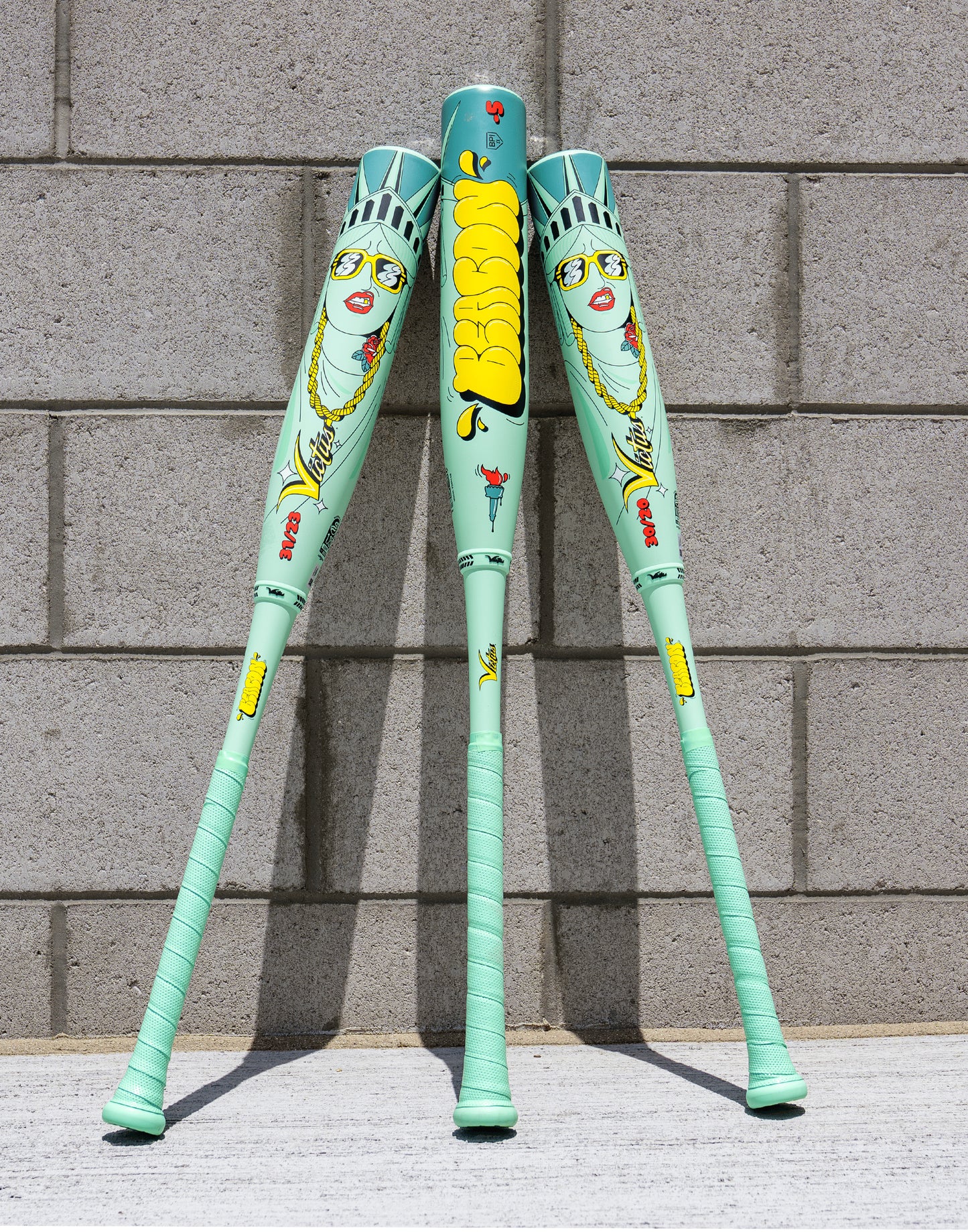 Three mint-green XL Victus 2027 Nova Lit Beacon (-10) USSSA baseball bats, featuring cartoon Statue of Liberty faces, yellow "DeeSon" text, carbon fiber details, and red/yellow graphics, stand against a gray brick wall.