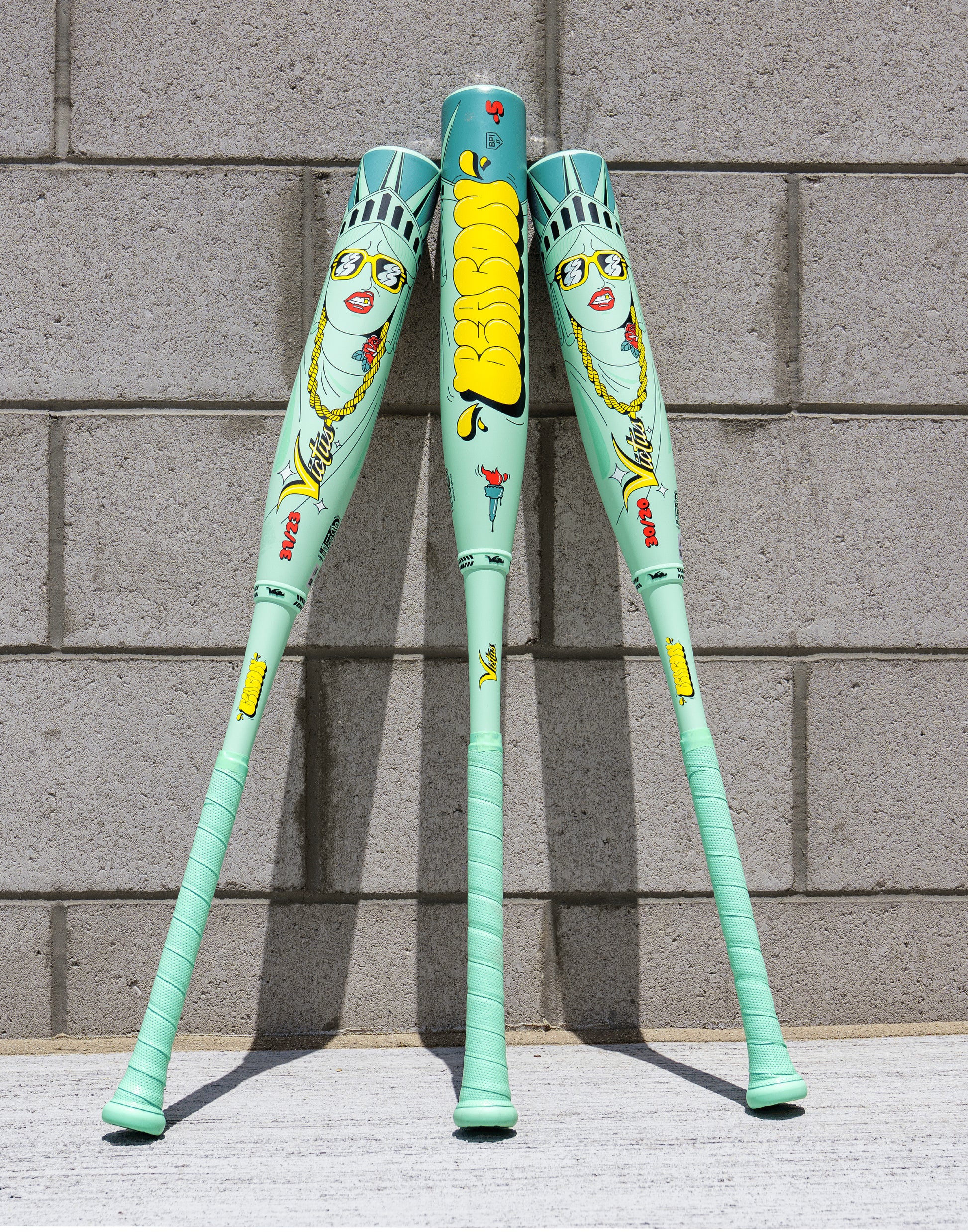 Three mint-green XL Victus 2027 Nova Lit Beacon (-10) USSSA baseball bats, featuring cartoon Statue of Liberty faces, yellow "DeeSon" text, carbon fiber details, and red/yellow graphics, stand against a gray brick wall.