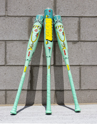 Three mint-green XL Victus 2027 Nova Lit Beacon (-10) USSSA baseball bats, featuring cartoon Statue of Liberty faces, yellow "DeeSon" text, carbon fiber details, and red/yellow graphics, stand against a gray brick wall.