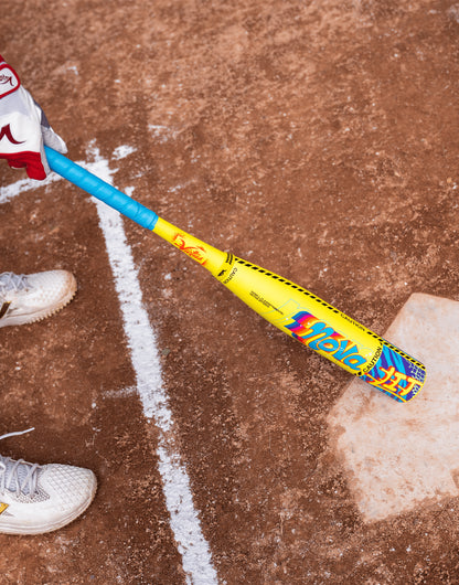 A person wearing white cleats and a red glove holds a 2026 Victus Nova Lit (-8) USSSA Baseball Bat (VSBNL8) from Victus, featuring yellow and blue colors, while standing by home plate on a dirt field.