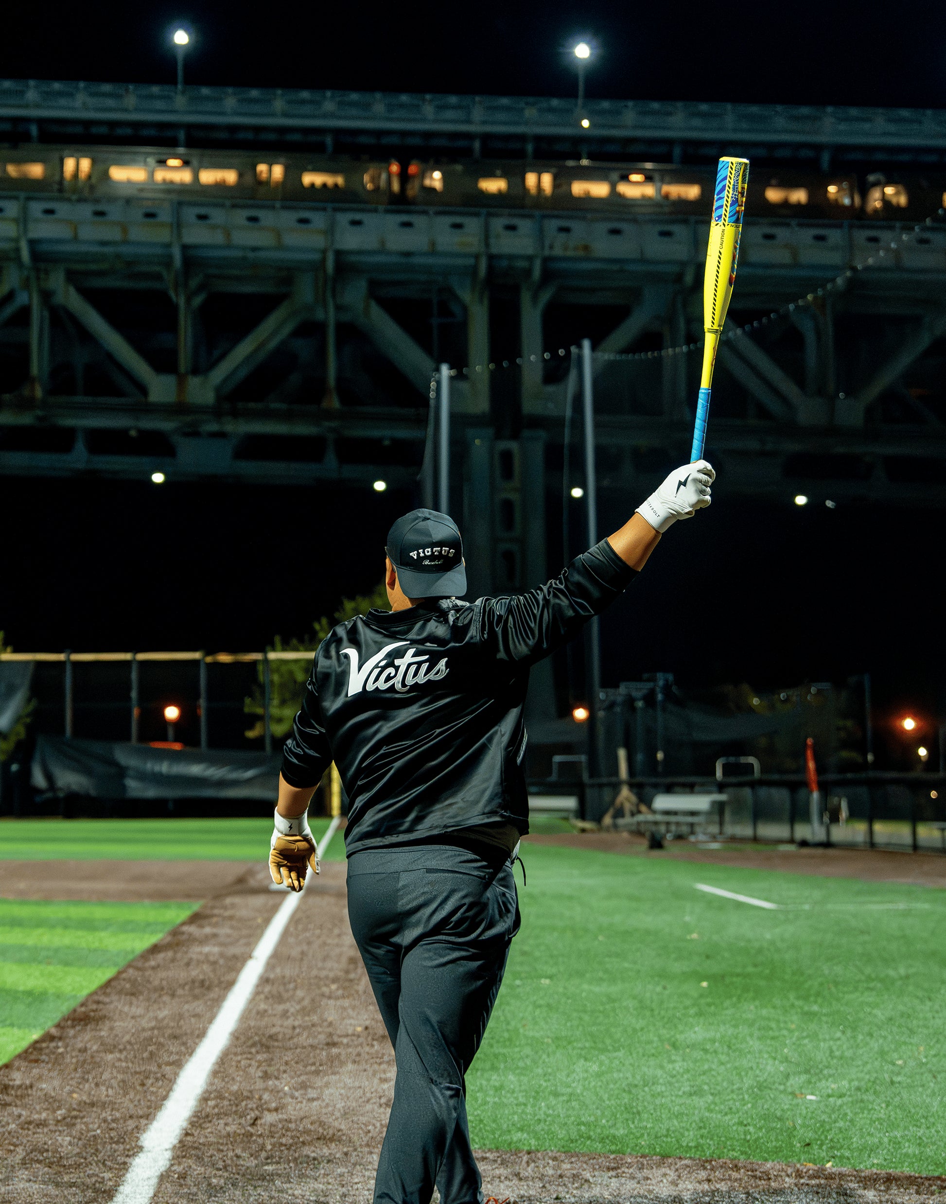 At night on the field, a baseball player in all black with “Victus” on his back raises the 2026 Victus Nova Lit (-10) USSSA Baseball Bat (VSBNL10) by Victus, while city lights and a glowing bridge shine in the background.