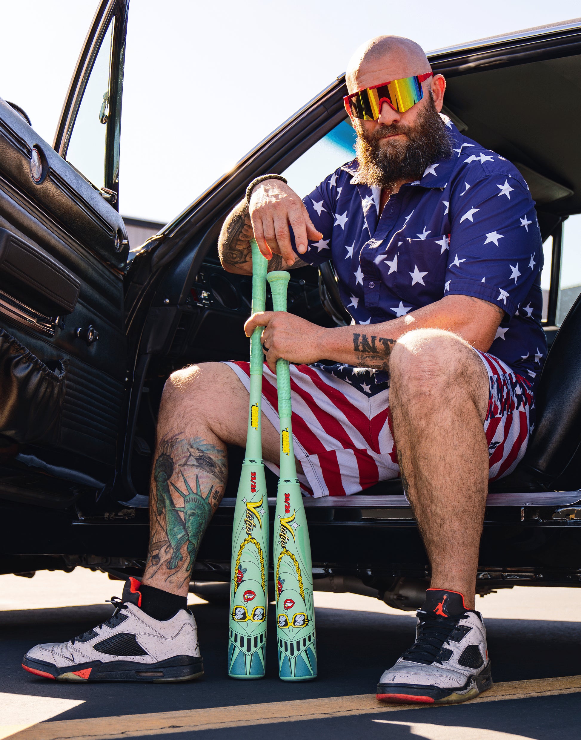 A bearded man in a star-patterned shirt, striped shorts, and colorful sunglasses sits in a car with the door open, holding two Victus Nova Lit Beacon (-8) baseball bats (VSBNL8B), showing off his tattoos and sneakers.