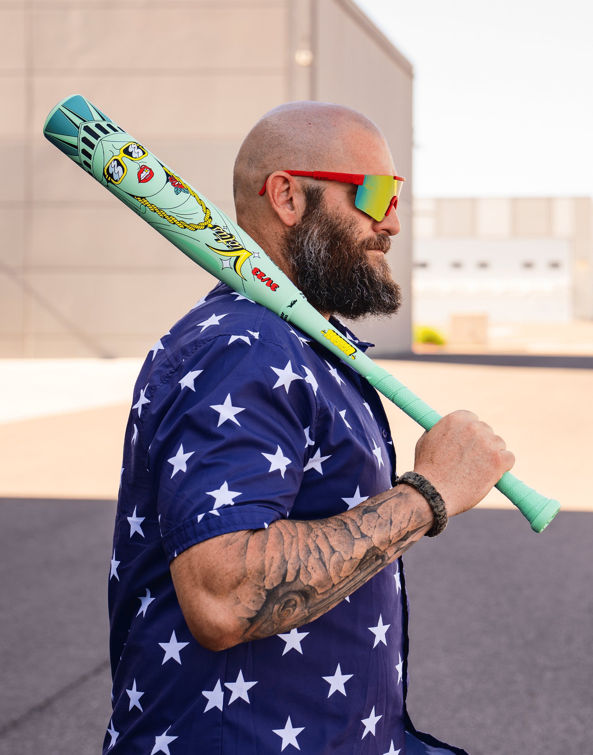 A bearded man in a navy blue shirt with white stars and red sunglasses holds a Victus 2027 Nova Lit Beacon (-10) USSSA Baseball Bat (VSBNL10B) featuring a cartoon Statue of Liberty, standing outdoors by a building.