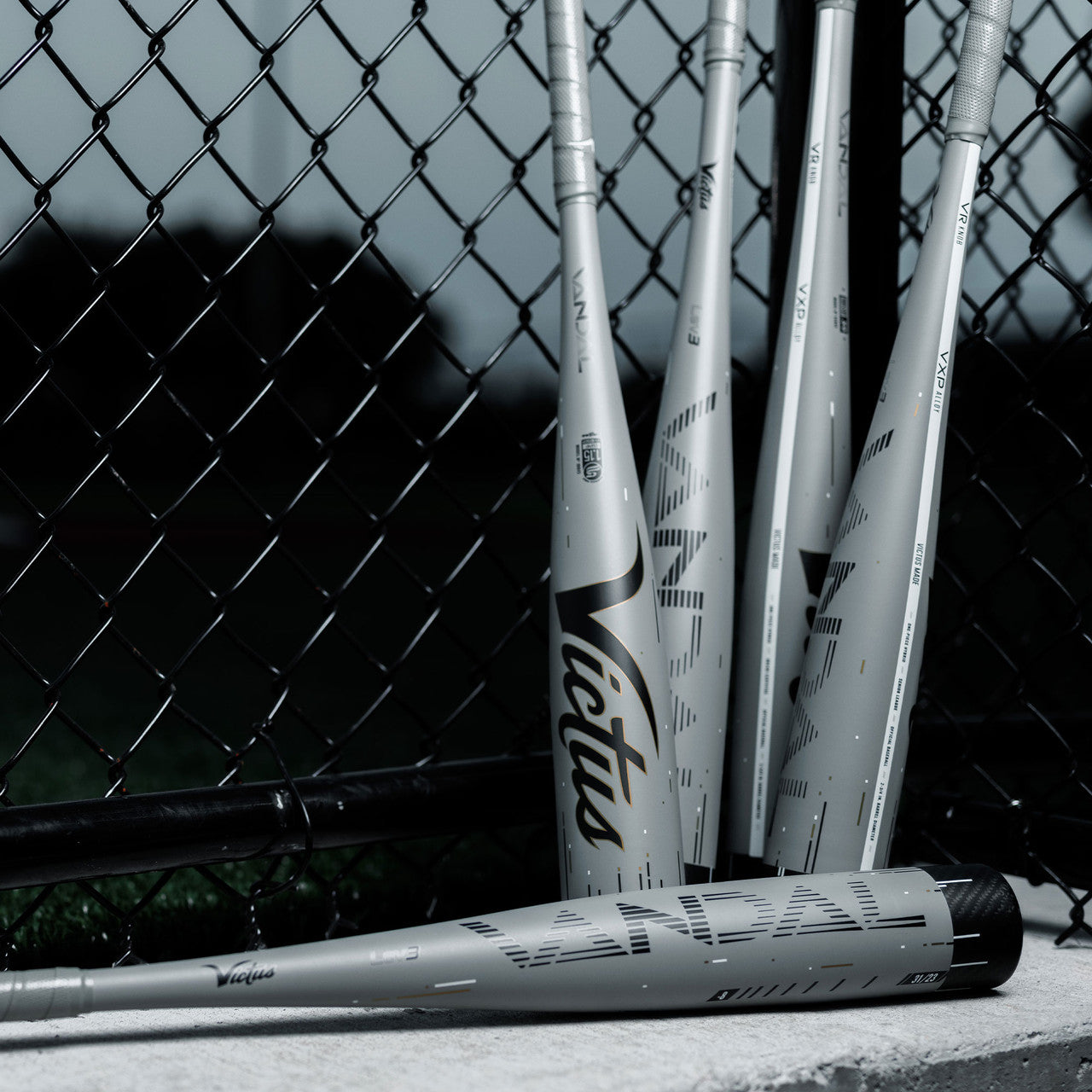 Four gray Victus Senior League bats, including a 2024 Victus Vandal Lev3 (-5) 2 3/4" USSSA Baseball Bat (USED), lean against a chain-link fence at a baseball field with grass in the background.