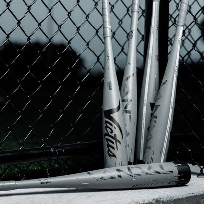 Four gray Victus Senior League bats, including a 2024 Victus Vandal Lev3 (-5) 2 3/4" USSSA Baseball Bat (USED), lean against a chain-link fence at a baseball field with grass in the background.