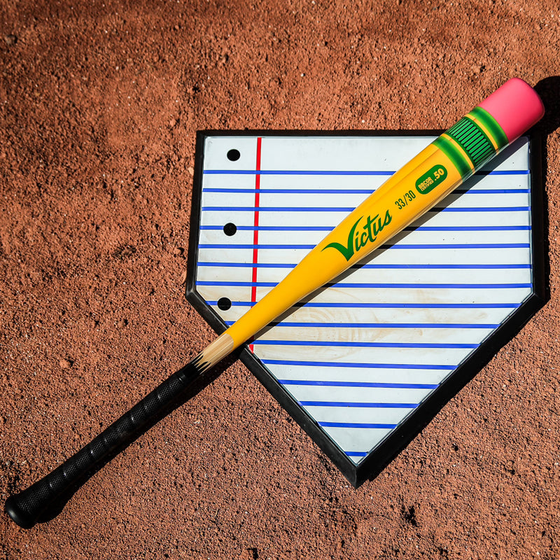 A yellow baseball bat designed to look like a pencil rests on a clipboard shaped like home plate with lined paper, set on a dirt baseball field.