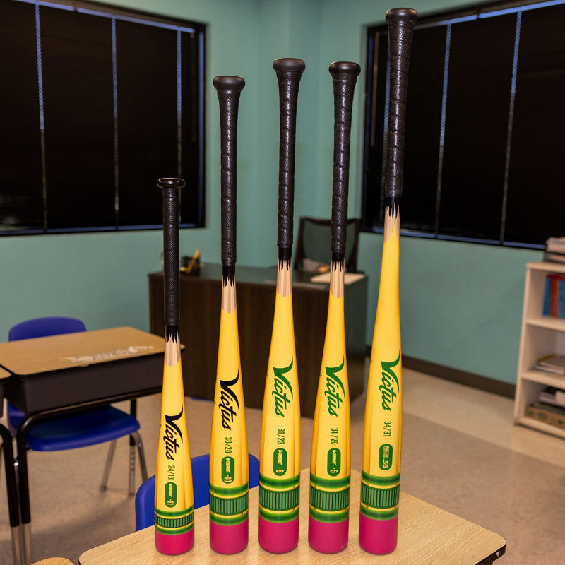 Five 2025 Victus Vibe Pencil (-5) bats by Victus—designed like yellow pencils with black eraser tips and green metal bands and made from extreme performance alloy—stand upright on and beside a classroom desk with blue chairs and black window blinds.
