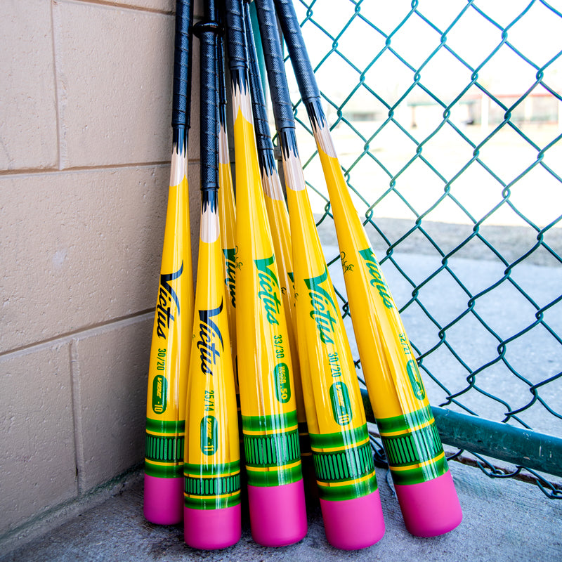 Six yellow baseball bats designed to look like pencils, with pink eraser-style ends and green/yellow details, lean against a beige brick wall near a chain-link fence on a sunny day.