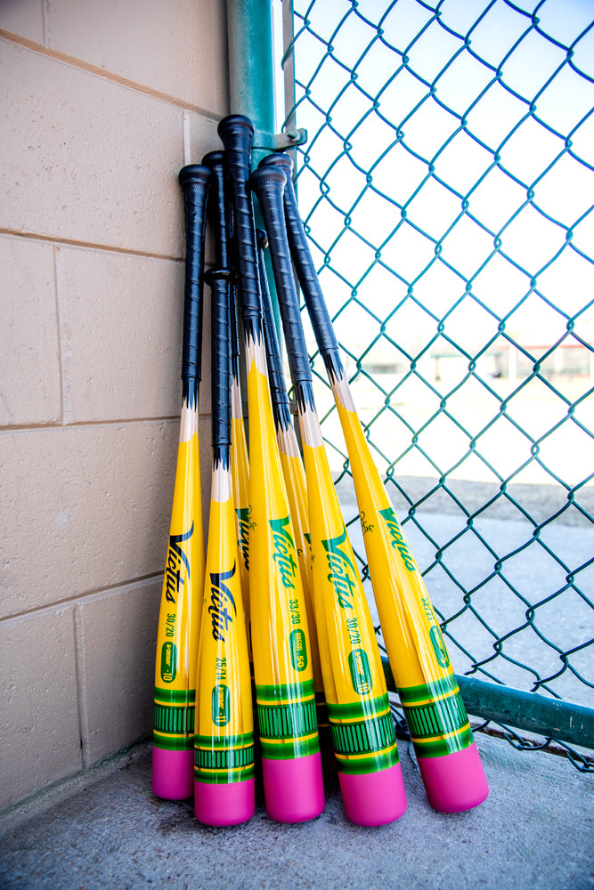 Seven yellow 2025 Victus Vibe Pencil (-5) USSSA Baseball Bats by Victus, with green and pink accents, are stacked together against a beige wall and chain-link fence at a sports field.
