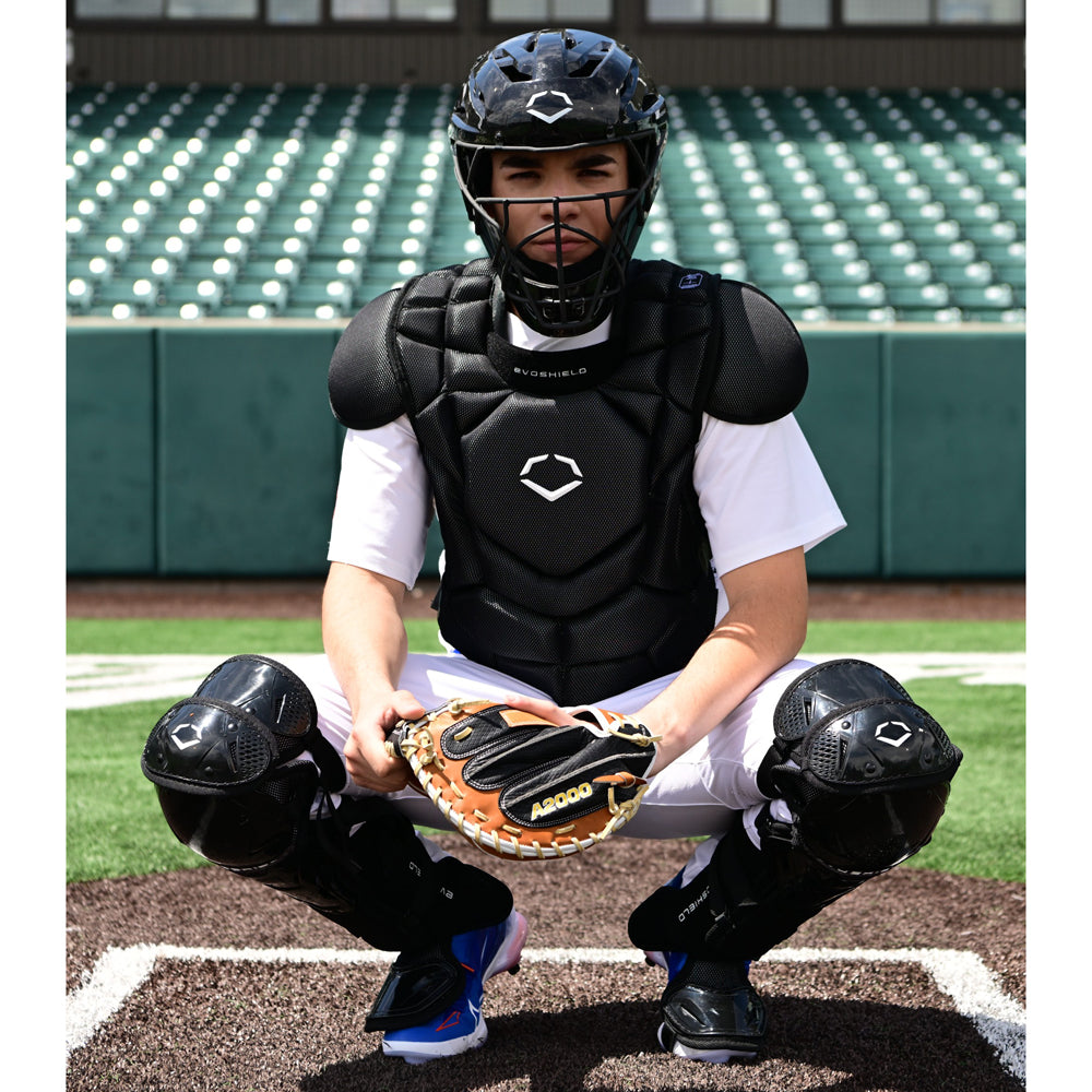 Wearing the EvoShield G2S Baseball Catcher's Gear Kit by EvoShield, a catcher crouches behind home plate with a mitt in an empty stadium.