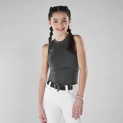 A young girl with dark braided hair smiles, wearing the EvoShield Women's Racerback Chest Guard Sleeveless Shirt (WB5747701/WB5747601) by EvoShield and white pants with a black belt, standing before a plain light gray background.