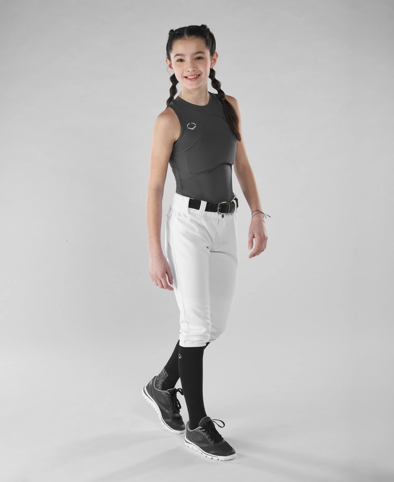 A young girl with braided hair smiles while wearing an EvoShield Women's Racerback Chest Guard Sleeveless Shirt, white baseball pants, black belt, socks, and sneakers, standing against a plain light gray background.