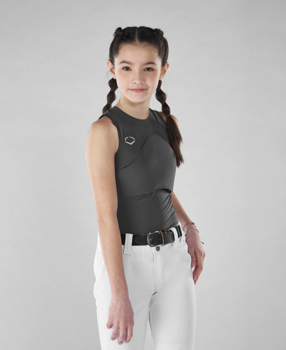 A young girl with braided hair stands against a gray background, wearing the EvoShield Women's Racerback Chest Guard Sleeveless Shirt (WB5747701/WB5747601) and white pants with a black belt. She smiles slightly, her right hand by her side.