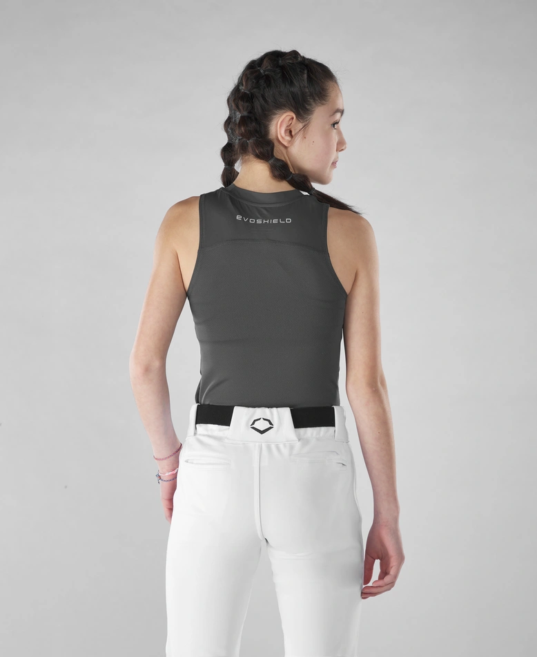 A young person with braided hair is seen from behind wearing an EvoShield Women's Racerback Chest Guard Sleeveless Shirt (WB5747701 / WB5747601), tucked into white athletic pants with a black belt against a plain gray background.
