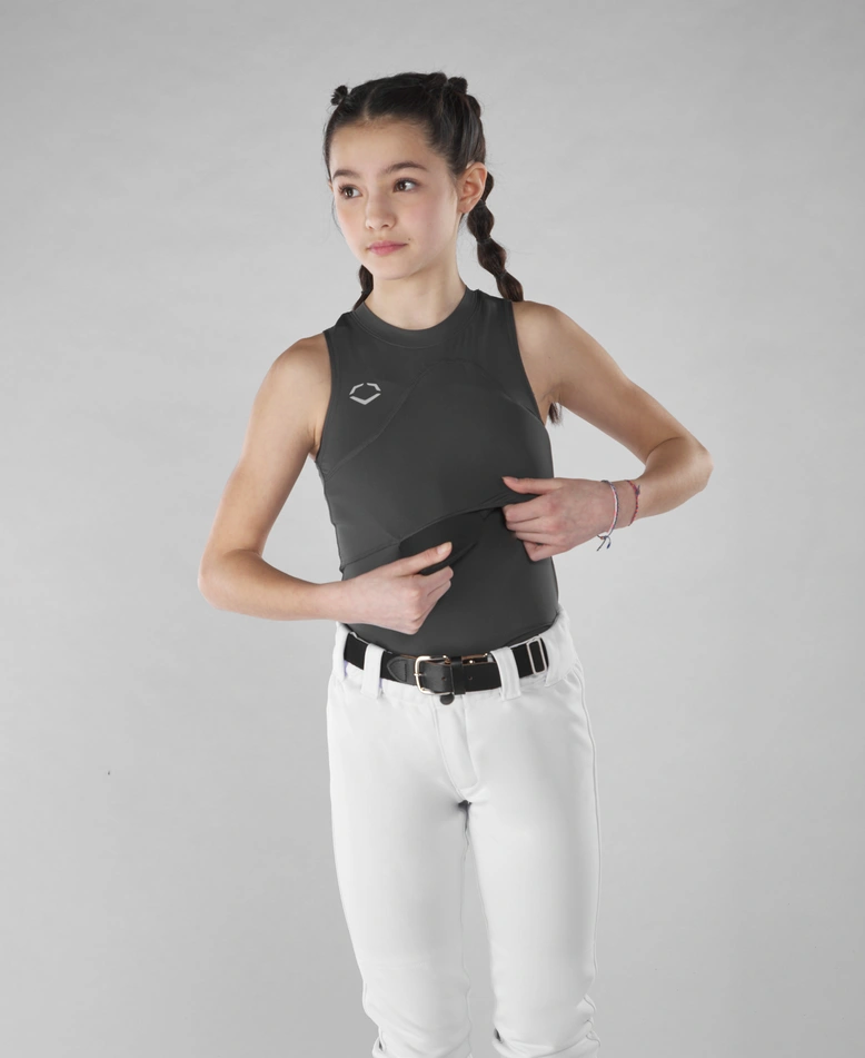 A young girl with braided hair models the EvoShield Women's Racerback Chest Guard Sleeveless Shirt (WB5747701/WB5747601) with white pants, standing against a plain gray background and looking slightly to the side.