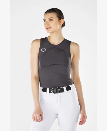 A woman wears the EvoShield Women's Racerback Chest Guard Sleeveless Shirt in dark gray with a white logo, paired with white pants and a black belt, standing against a light background and smiling slightly, looking off to the side.
