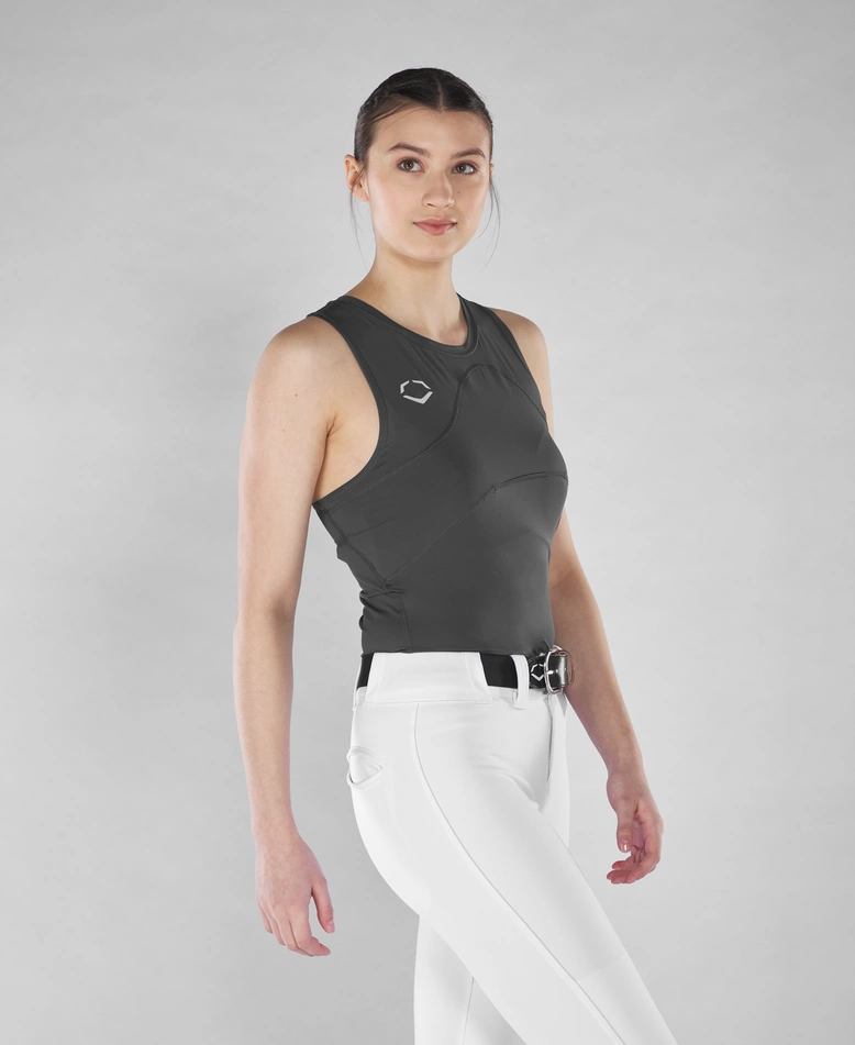 A woman with dark hair in a low ponytail models the EvoShield Women's Racerback Chest Guard Sleeveless Shirt, featuring a small chest logo, paired with high-waisted white pants and a black belt against a light gray background.